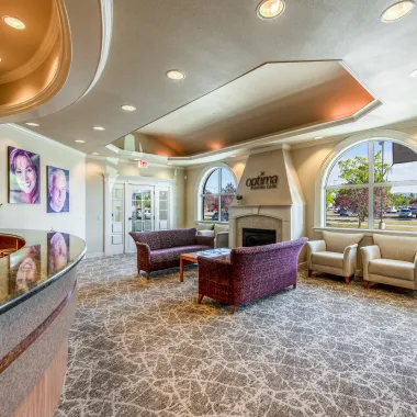 Reception desk area with a modern curved design, open ceiling detail, and decorative plants, adjacent to the patient waiting area with purple sofas.